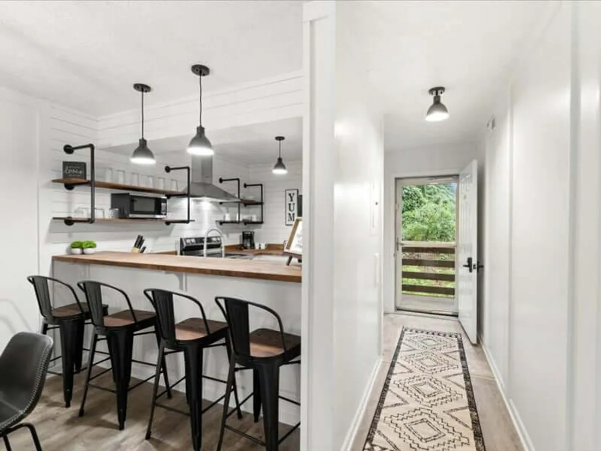 A bright, modern kitchen featuring a kitchen island with bar stools, sleek cabinetry, and decorative wall art. The space flows seamlessly to an open outdoor area visible through the door, emphasizing both functionality and a social, inviting atmosphere.