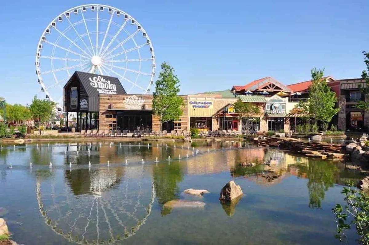 The scene shows a lively outdoor setting with a ferris wheel, pond, quaint wooden buildings, and bustling shops. People can be seen walking, shopping, and enjoying the vibrant atmosphere.