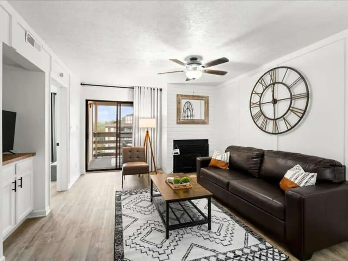 This image features a large wall clock, a brown leather sofa, a coffee table with decorative items, and a modern light fixture. The space is styled with a mix of contemporary décor and cozy details, promoting comfort and a welcoming environment. The ample floor space and open arrangement suggest an open-plan layout.