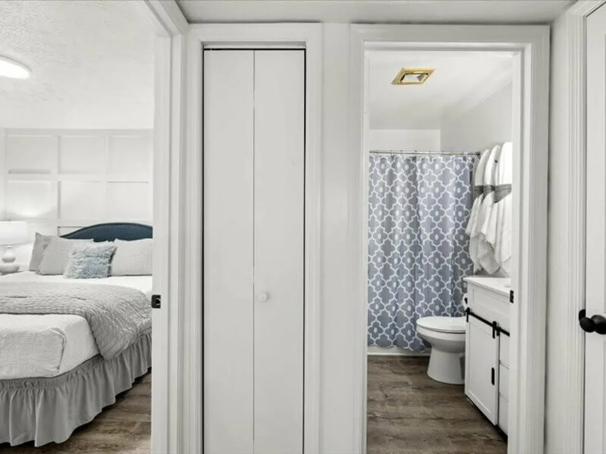 A well-organized bedroom with a neatly made bed, a bathroom visible with a patterned shower curtain, and tidy closet space. The room exudes a serene, organized feel, ideal for relaxation and refreshment.