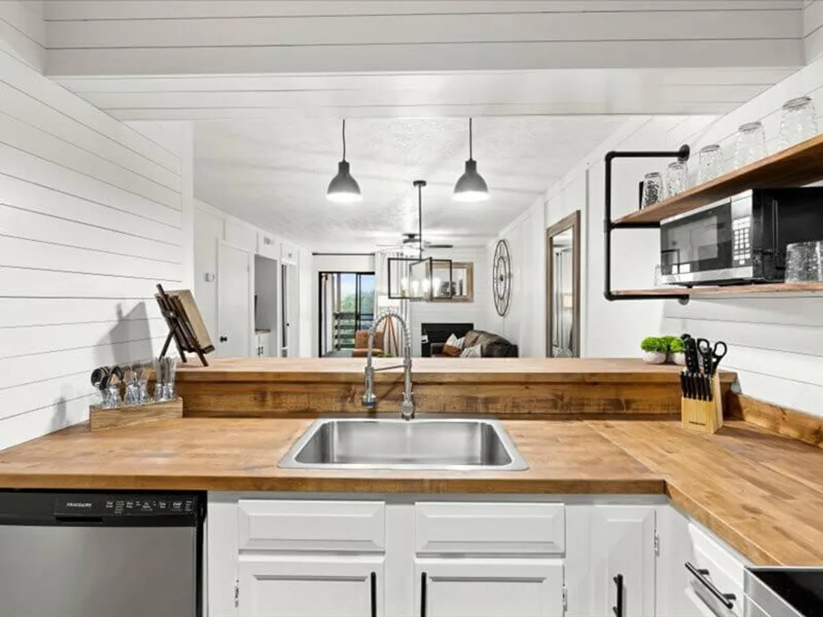 A cozy kitchen space featuring a wooden countertop, open shelving, pendant lights, and a spacious sink area. The ambiance is warm and perfect for gatherings or casual meal prep.