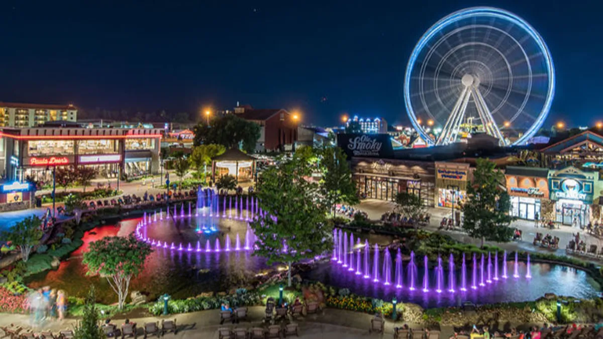 A lively downtown strip features a Ferris wheel, a colorful fountain, shops, restaurants, and people enjoying the space. The energetic atmosphere is perfect for couples strolling hand-in-hand, discovering wine bars, boutiques, and local dining options.
