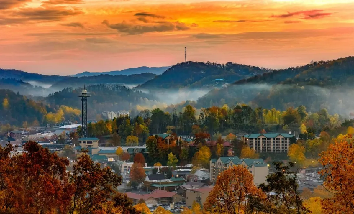 The image captures a sweeping view of distant mountains, a small town below, and a glowing sunset sky with vibrant fall foliage in the foreground. The tranquil scene invites exploration and embodies the beauty of Gatlinburg’s surroundings.