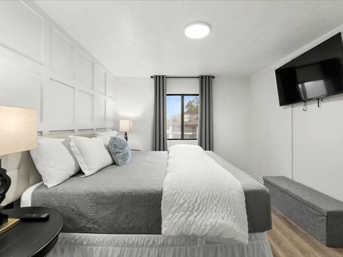 A tranquil bedroom scene showing a bed dressed in grey and white bedding, a decorative pillow, a nightstand with a lamp, and a wall-mounted television. The room is brightened by natural light, combining modern comfort with calm aesthetics.