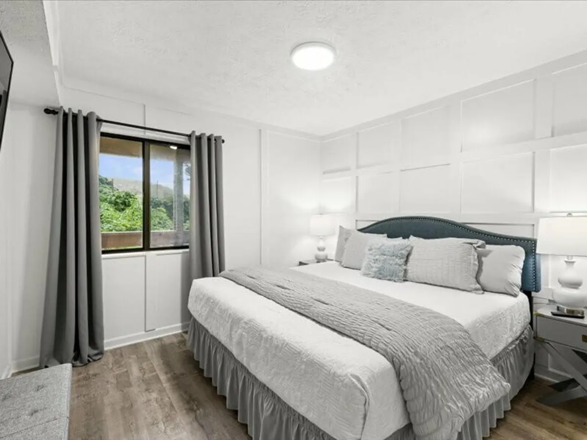 A serene bedroom featuring a comfortable bed with decorative pillows, a large window with flowing curtains offering views of the outdoors, a nightstand with a lamp, and subtle modern decor. The natural light fills the room, enhancing its tranquil and inviting atmosphere.