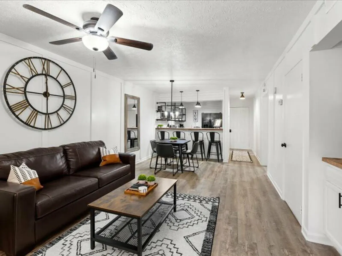 An inviting open-plan living area featuring a brown leather sofa, a statement wall clock, a dining table with sleek black chairs, and a geometric area rug. Natural light floods the room, highlighting both the modern furnishings and the spacious layout ideal for socializing.